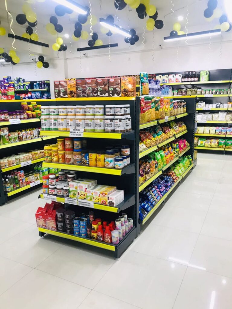 Display Racks for Shop in Dehradun Shelving & rack manufacturer in Ludhiana: Multiple rows of black shelves with bright yellow edges, stocked with various food products. Products: A wide variety of cereals, including recognizable brands like Kellogg's Many types of snacks, particularly corn-based snacks like tortilla chips Different flavors and brands of chips and crunchy snacks Some oatmeal and breakfast products Organization: The products are neatly arranged and grouped by type and brand. Signage: There's a small sign visible that says "CEREALS" indicating the product category. Decoration: The ceiling is decorated with balloons in black and yellow colors, along with some streamers. This suggests the store may be celebrating an opening, anniversary, or special event. Lighting: Bright fluorescent lights hang from the ceiling, providing ample illumination for the products. Layout: The shelves are arranged in an L-shape, with the corner of the aisle visible in the image.