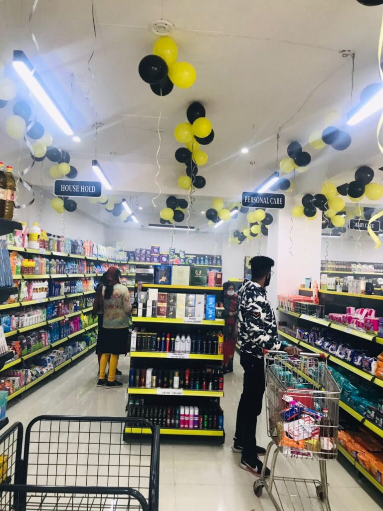 Display Racks for Shop in Dehradun Shelving: Multiple rows of black shelves with bright yellow edges, stocked with various food products. Products: A wide variety of cereals, including recognizable brands like Kellogg's Many types of snacks, particularly corn-based snacks like tortilla chips Different flavors and brands of chips and crunchy snacks Some oatmeal and breakfast products Organization: The products are neatly arranged and grouped by type and brand. Signage: There's a small sign visible that says "CEREALS" indicating the product category. Decoration: The ceiling is decorated with balloons in black and yellow colors, along with some streamers. This suggests the store may be celebrating an opening, anniversary, or special event. Lighting: Bright fluorescent lights hang from the ceiling, providing ample illumination for the products. Layout: The shelves are arranged in an L-shape, with the corner of the aisle visible in the image.