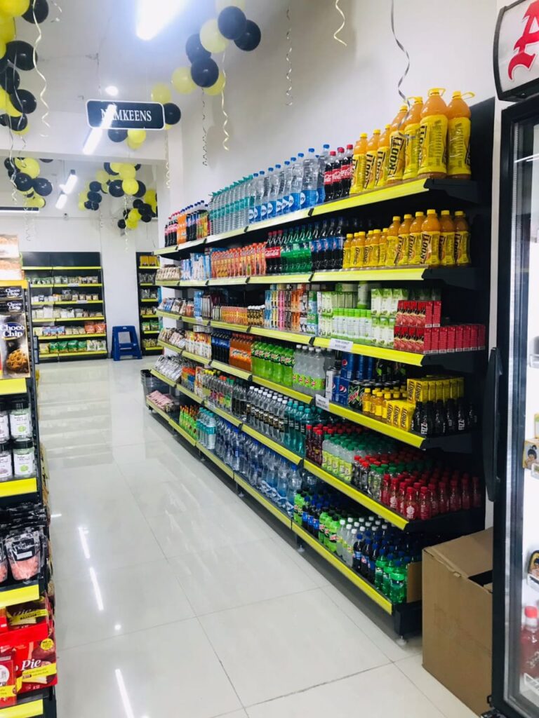 Display Racks for Shop in Dehradun Shelving: Multiple rows of black shelves with bright yellow edges, stocked with various food products. Products: A wide variety of cereals, including recognizable brands like Kellogg's Many types of snacks, particularly corn-based snacks like tortilla chips Different flavors and brands of chips and crunchy snacks Some oatmeal and breakfast products Organization: The products are neatly arranged and grouped by type and brand. Signage: There's a small sign visible that says "CEREALS" indicating the product category. Decoration: The ceiling is decorated with balloons in black and yellow colors, along with some streamers. This suggests the store may be celebrating an opening, anniversary, or special event. Lighting: Bright fluorescent lights hang from the ceiling, providing ample illumination for the products. Layout: The shelves are arranged in an L-shape, with the corner of the aisle visible in the image.