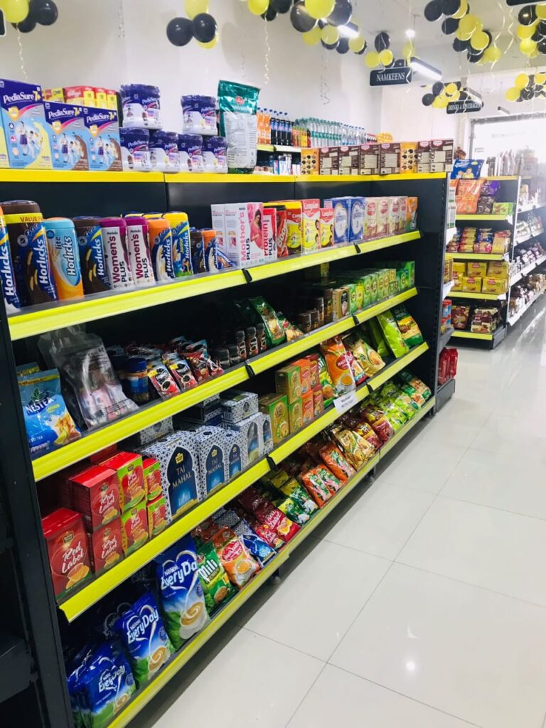 Display Racks for Shop in Dehradun Shelving: Multiple rows of black shelves with bright yellow edges, stocked with various food products. Products: A wide variety of cereals, including recognizable brands like Kellogg's Many types of snacks, particularly corn-based snacks like tortilla chips Different flavors and brands of chips and crunchy snacks Some oatmeal and breakfast products Organization: The products are neatly arranged and grouped by type and brand. Signage: There's a small sign visible that says "CEREALS" indicating the product category. Decoration: The ceiling is decorated with balloons in black and yellow colors, along with some streamers. This suggests the store may be celebrating an opening, anniversary, or special event. Lighting: Bright fluorescent lights hang from the ceiling, providing ample illumination for the products. Layout: The shelves are arranged in an L-shape, with the corner of the aisle visible in the image.