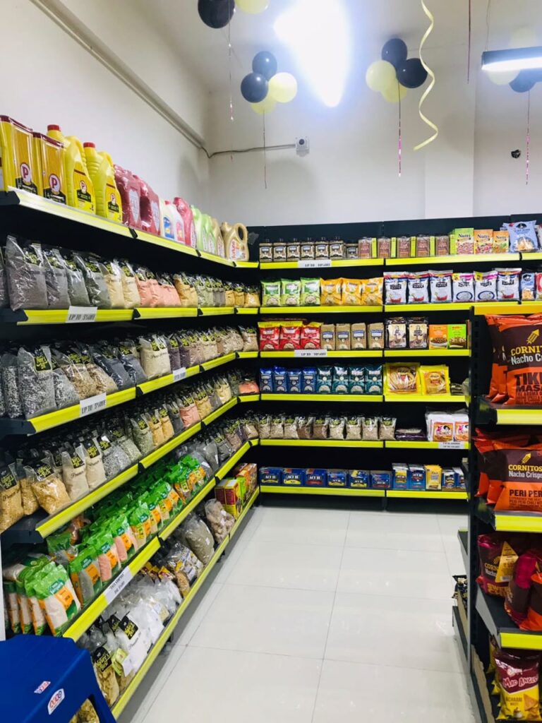 Display Racks for Shop in Dehradun Shelving & rack manufacturer in Ludhiana: Multiple rows of black shelves with bright yellow edges, stocked with various food products. Products: A wide variety of cereals, including recognizable brands like Kellogg's Many types of snacks, particularly corn-based snacks like tortilla chips Different flavors and brands of chips and crunchy snacks Some oatmeal and breakfast products Organization: The products are neatly arranged and grouped by type and brand. Signage: There's a small sign visible that says "CEREALS" indicating the product category. Decoration: The ceiling is decorated with balloons in black and yellow colors, along with some streamers. This suggests the store may be celebrating an opening, anniversary, or special event. Lighting: Bright fluorescent lights hang from the ceiling, providing ample illumination for the products. Layout: The shelves are arranged in an L-shape, with the corner of the aisle visible in the image.