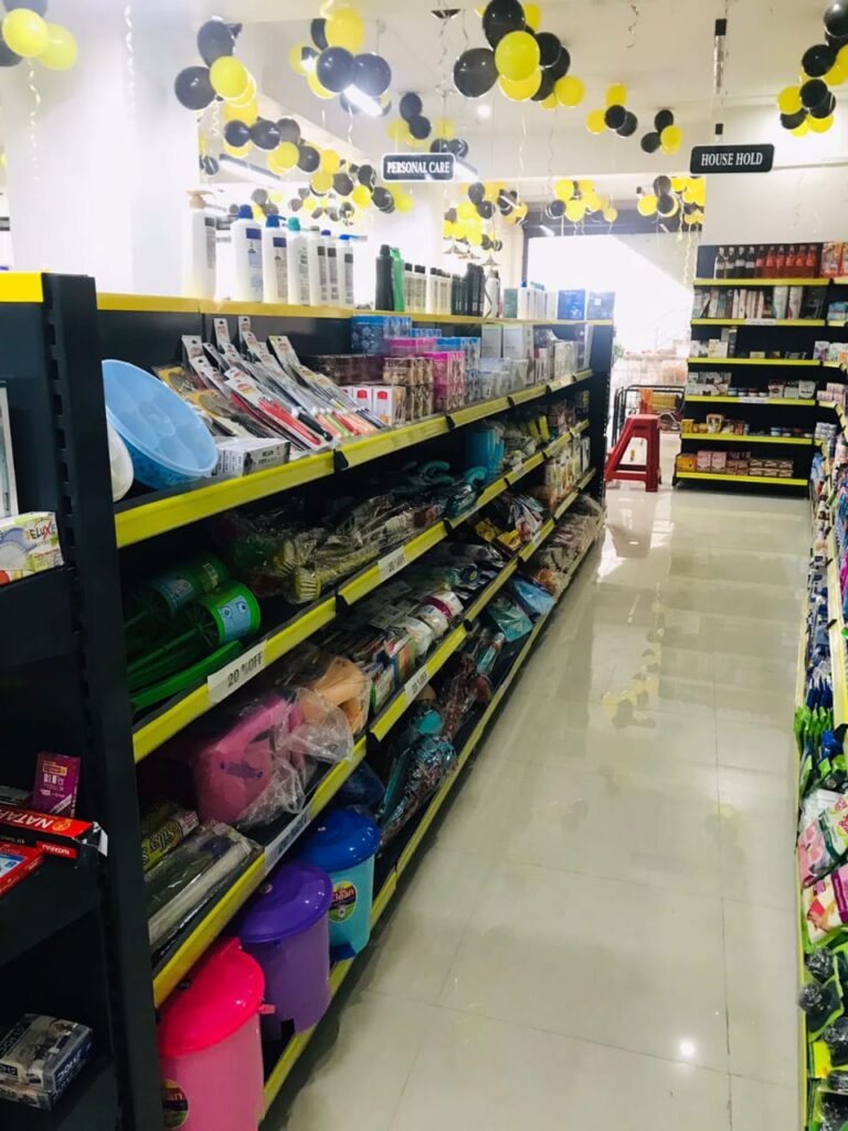 Display Racks for Shop in Dehradun Shelving & rack manufacturer in Ludhiana: Multiple rows of black shelves with bright yellow edges, stocked with various food products. Products: A wide variety of cereals, including recognizable brands like Kellogg's Many types of snacks, particularly corn-based snacks like tortilla chips Different flavors and brands of chips and crunchy snacks Some oatmeal and breakfast products Organization: The products are neatly arranged and grouped by type and brand. Signage: There's a small sign visible that says "CEREALS" indicating the product category. Decoration: The ceiling is decorated with balloons in black and yellow colors, along with some streamers. This suggests the store may be celebrating an opening, anniversary, or special event. Lighting: Bright fluorescent lights hang from the ceiling, providing ample illumination for the products. Layout: The shelves are arranged in an L-shape, with the corner of the aisle visible in the image.