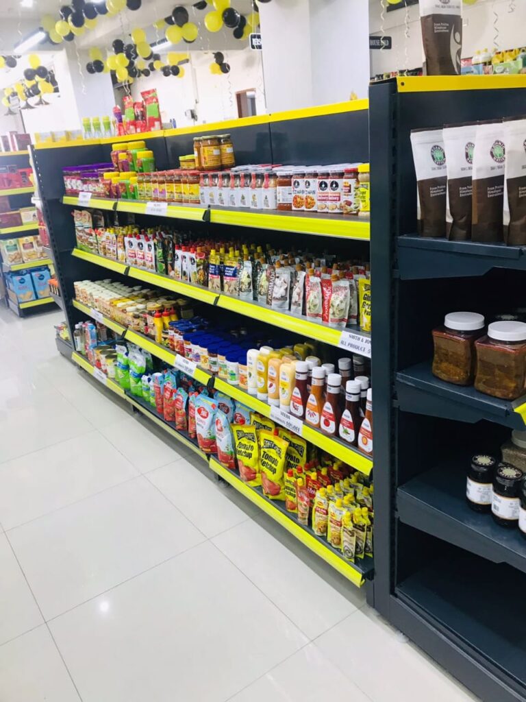 Display Racks for Shop in Dehradun Shelving & rack manufacturer in Ludhiana: Multiple rows of black shelves with bright yellow edges, stocked with various food products. Products: A wide variety of cereals, including recognizable brands like Kellogg's Many types of snacks, particularly corn-based snacks like tortilla chips Different flavors and brands of chips and crunchy snacks Some oatmeal and breakfast products Organization: The products are neatly arranged and grouped by type and brand. Signage: There's a small sign visible that says "CEREALS" indicating the product category. Decoration: The ceiling is decorated with balloons in black and yellow colors, along with some streamers. This suggests the store may be celebrating an opening, anniversary, or special event. Lighting: Bright fluorescent lights hang from the ceiling, providing ample illumination for the products. Layout: The shelves are arranged in an L-shape, with the corner of the aisle visible in the image.