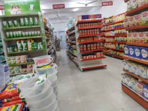 (Dukan Ke Racks) kirana store rack in Bhopal, Madhya Pradesh. Racks Manufactured by GKSP RACKING. This image shows a different section of the store. On the left side, there's a green display stand with skincare products, advertising "Clinically Proven Pimple-free Healthy skin". The center aisle contains various food products, and on the right, there are more packaged foods. The Front gaurd of the shelve is red.