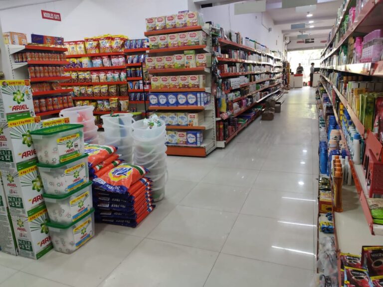 (Dukan Ke Racks) kirana store rack in Bhopal, Madhya Pradesh. Racks Manufactured by GKSP RACKING. This image shows another view of the same store aisle. It's wider than the first image and includes more variety of products. On the left side, there are stacks of Ariel detergent boxes and plastic storage containers. The right side shows shelves stocked with various packaged foods, including breakfast cereals. There's a sign visible that says "NOODLES/OATS".