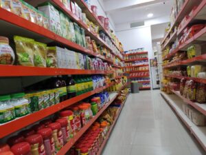 (Dukan Ke Racks) kirana store rack in Bhopal, Madhya Pradesh. Racks Manufactured by GKSP RACKING. This image shows an aisle in a grocery store/supermarket. The front gaurd of shelves are red and stocked with various food items and household products. The aisle well-organized and brightly lit. Products visible include packaged foods, beverages, and cooking oils/sauces on the lower shelves.