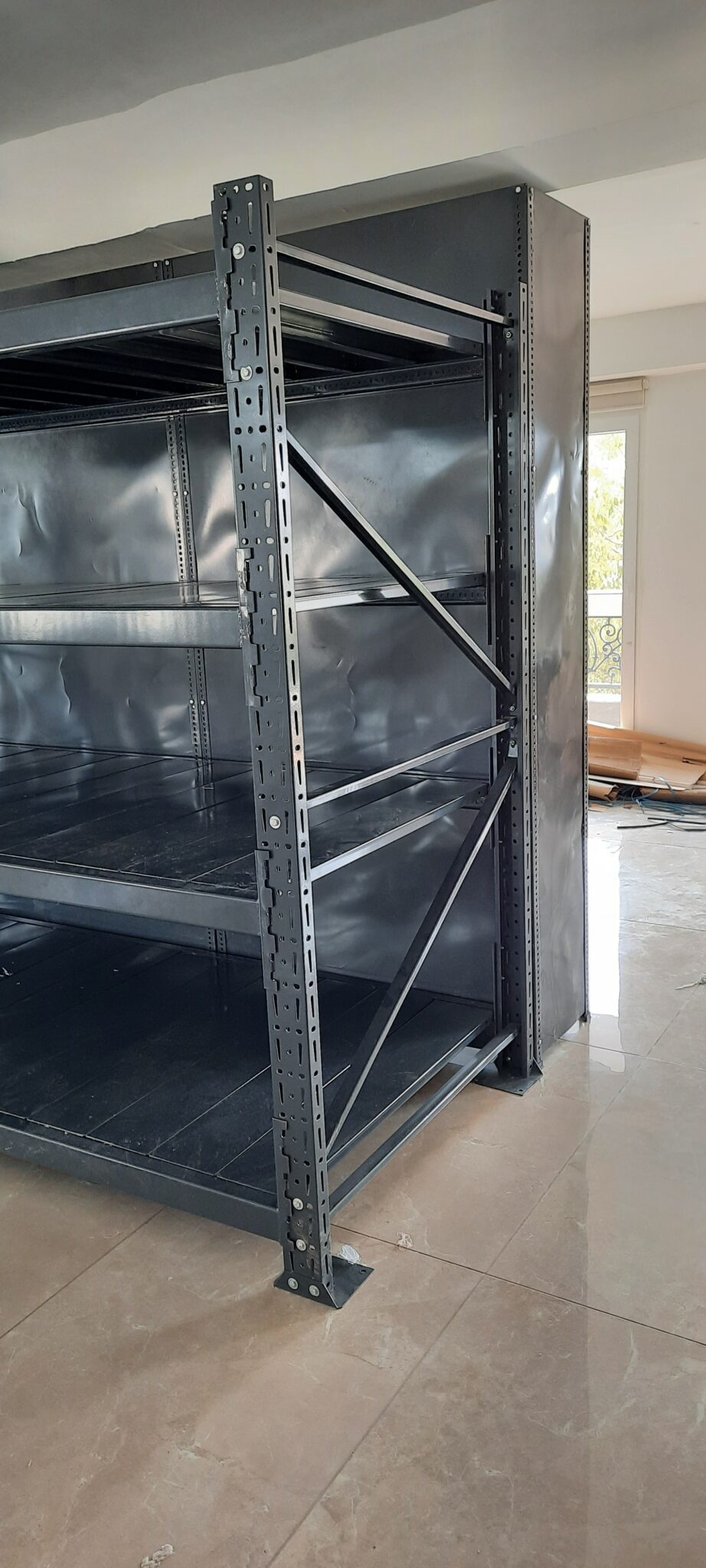 This shows a close-up view of an industrial-style heavy duty pallet rack in Uttarakhand. It's made of dark gray with multiple shelf levels. The upright support post is with many regular holes for adjustable shelf heights. The unit is installed on a tiled floor in a room with white walls.