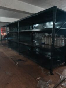 This is a darker shot of what appears to be the heavy duty pallet rack in Uttarakhand, shelving system, but with some items stored on it, including large water jugs or containers. The shelving unit extends quite long and has side panels installed. The room is under construction with some debris on the wooden floor.