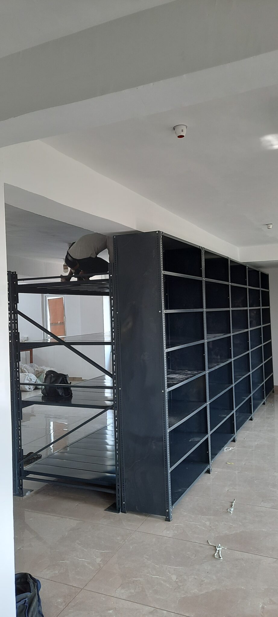 This is a full-height view of the completed shelving unit or the heavy duty pallet rack and the slotted angle rack in Uttarakhand installation. A person can is working on top of the unit, doing installation or adjustment work. The unit is 7 feet tall, reaching nearly to the ceiling, and has multiple sections side by side creating a wall of shelving. The room has modern lighting fixtures including smoke detector on the ceiling. The flooring is finished tile and the walls are painted white.