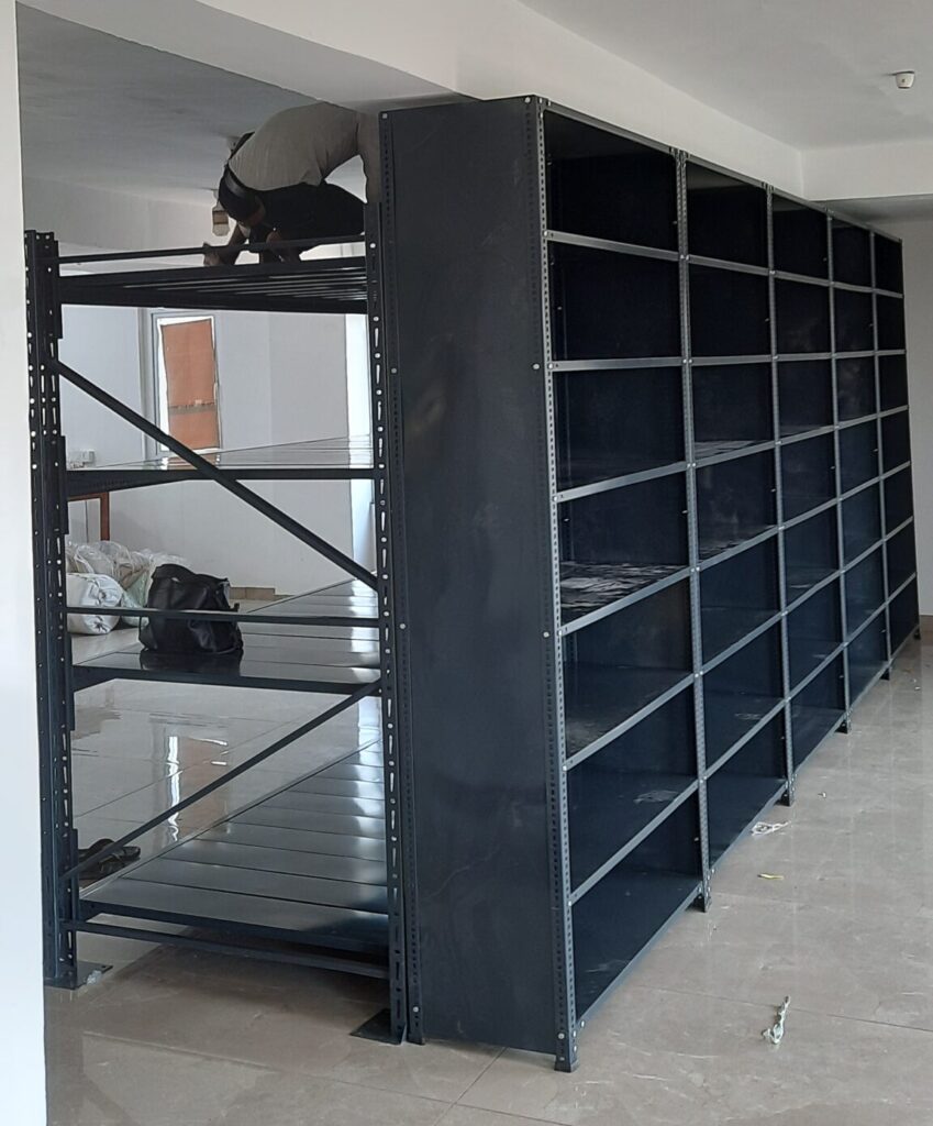 This is a full-height view of the completed shelving unit or the heavy duty pallet rack and the slotted angle rack in Uttarakhand installation. A person can is working on top of the unit, doing installation or adjustment work. The unit is 7 feet tall, reaching nearly to the ceiling, and has multiple sections side by side creating a wall of shelving. The room has modern lighting fixtures including smoke detector on the ceiling. The flooring is finished tile and the walls are painted white.