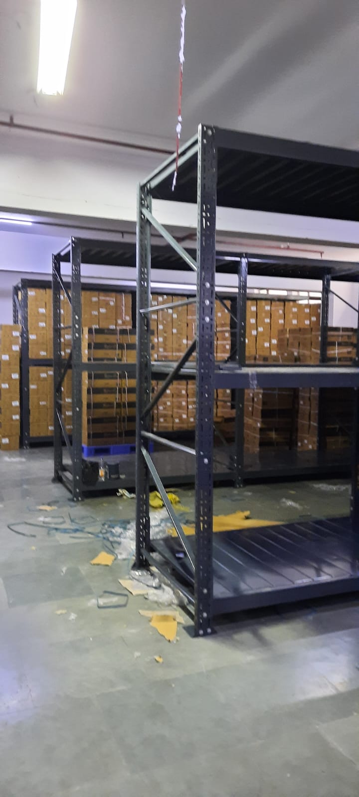 This image depicts a warehouse storage area in the process of installing heavyduty pallet rack. Heavy Duty Pallet Rack with Multi-level steel racking systems, dark gray or black in color, are visible. These racks are partially assembled and extend from floor to ceiling. Inventory Storage on The upper levels of the racks are filled with brown cardboard boxes, neatly stacked and labeled, suggesting organized inventory management. A white ceiling with visible fixtures and a red stripe, likely for utilities or building design. Concrete flooring with visible dust, debris, and materials scattered about. The racking system demonstrates efficient use of vertical space for high-density storage.