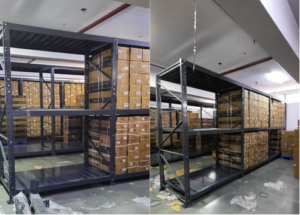 This image depicts a warehouse storage area in the process of installing heavyduty pallet rack. Heavy Duty Pallet Rack with Multi-level steel racking systems, dark gray or black in color, are visible. These racks are partially assembled and extend from floor to ceiling. Inventory Storage on The upper levels of the racks are filled with brown cardboard boxes, neatly stacked and labeled, suggesting organized inventory management. A white ceiling with visible fixtures and a red stripe, likely for utilities or building design. Concrete flooring with visible dust, debris, and materials scattered about. The racking system demonstrates efficient use of vertical space for high-density storage.