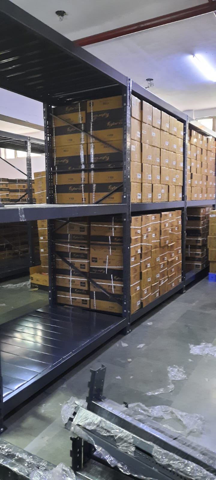 This image depicts a warehouse storage area in the process of installing heavyduty pallet rack. Heavy Duty Pallet Rack with Multi-level steel racking systems, dark gray or black in color, are visible. These racks are partially assembled and extend from floor to ceiling. Inventory Storage on The upper levels of the racks are filled with brown cardboard boxes, neatly stacked and labeled, suggesting organized inventory management. A white ceiling with visible fixtures and a red stripe, likely for utilities or building design. Concrete flooring with visible dust, debris, and materials scattered about. The racking system demonstrates efficient use of vertical space for high-density storage.