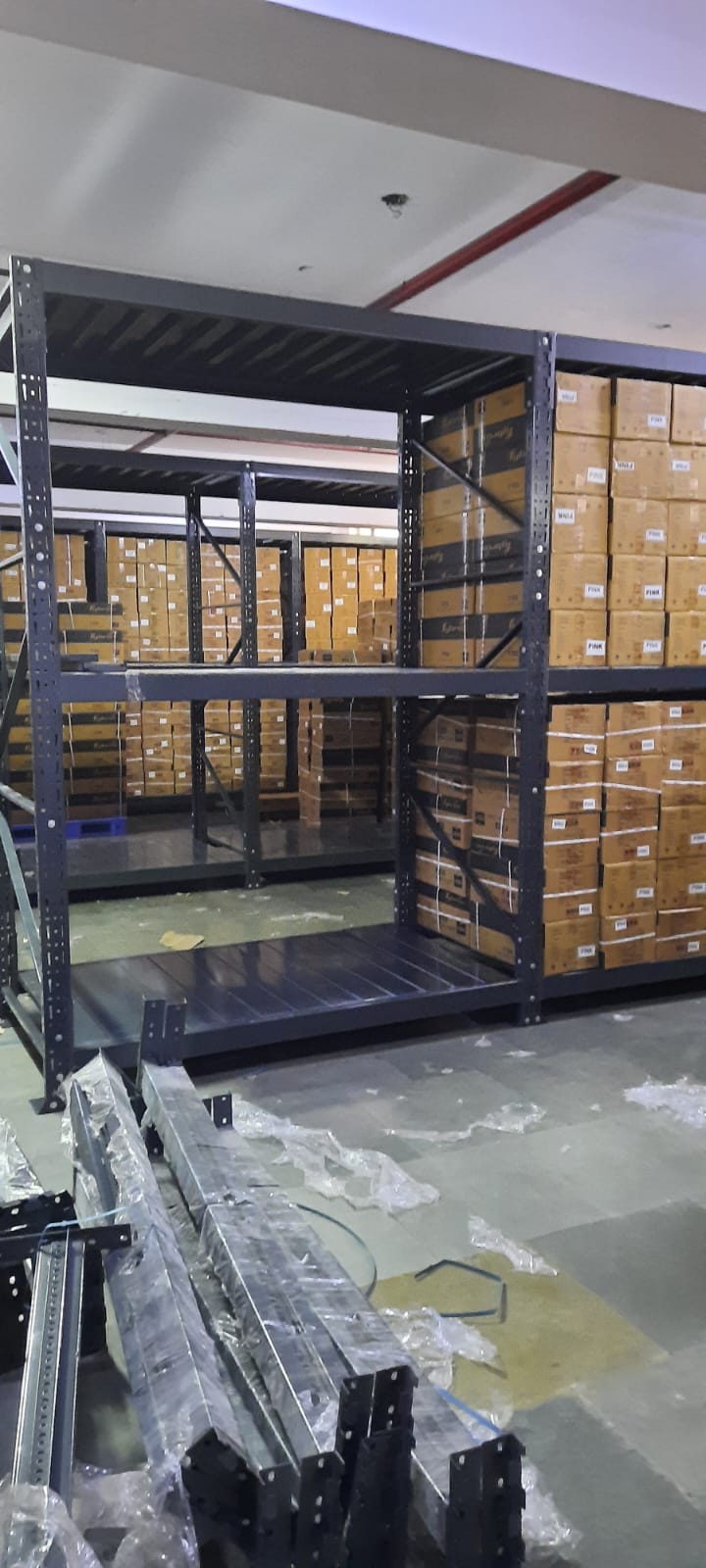 This image depicts a warehouse storage area in the process of installing heavyduty pallet rack. Heavy Duty Pallet Rack with Multi-level steel racking systems, dark gray or black in color, are visible. These racks are partially assembled and extend from floor to ceiling. Inventory Storage on The upper levels of the racks are filled with brown cardboard boxes, neatly stacked and labeled, suggesting organized inventory management. A white ceiling with visible fixtures and a red stripe, likely for utilities or building design. Concrete flooring with visible dust, debris, and materials scattered about. The racking system demonstrates efficient use of vertical space for high-density storage.